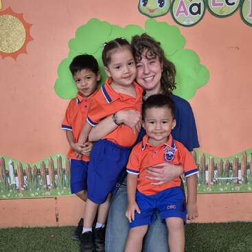 Eine lächelnde Lehrerin umarmt drei kleine Kinder in einheitlichen orange-blauen Uniformen. Sie posieren vor einer bunten Klassenzimmerwand, die mit Bäumen, einer Sonne und Buchstaben verziert ist.
