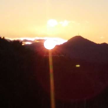 The image depicts a serene sunset scene, with the sun positioned behind a mountain, casting a warm glow on the sky and landscape.