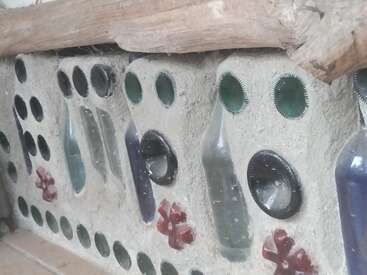 Dieses Bild zeigt eine Wand aus Glasflaschen, die in Beton eingebettet und mit Mustern verziert sind. Darauf liegen Holzbalken, die einen rustikalen, künstlerischen Touch verleihen.