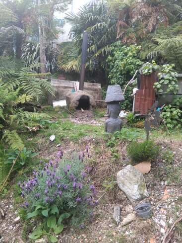 Eine üppige Gartenszene mit Farnen, Lavendel, einem rustikalen Outdoor-Ofen, Steinskulpturen, rostigen Blechen und verstreuten Steinen, die eine friedliche, eklektische Naturkulisse schaffen.