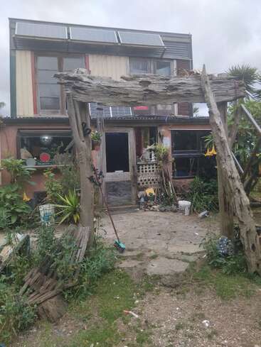 Ein rustikales, zweistöckiges Haus mit Sonnenkollektoren, einem Torbogen aus verwittertem Holz, einem überwucherten Garten, verstreuten Gegenständen und einem Besen. Die Atmosphäre ist schrullig und heimatverbunden.