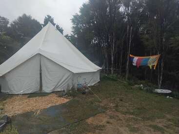 Ein großes weißes Zelt steht auf einem grasbewachsenen Boden in der Nähe eines Waldes. Bunte Gebetsfahnen hängen an einer Leine. Die Atmosphäre ist neblig und heiter.