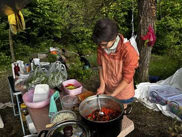 A person in an orange jacket prepares food outdoors, surrounded by vegetables and cooking supplies on a table, with a forest background and camping gear nearby.