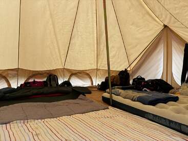 Inside a spacious, cream-colored tent, sleeping bags, air mattresses, and blankets are neatly arranged on the floor; backpacks and personal belongings line the tent’s edges.
