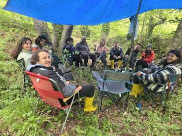 A group of people sit in a circle on camping chairs under a blue tarp, enjoying nature in a lush green forest, all wearing yellow boots.