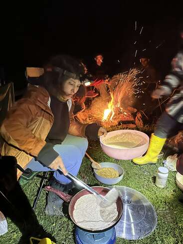 Una mujer, con una linterna frontal, prepara pan plano en un hornillo de camping por la noche, mientras otras personas se sientan alrededor de una hoguera, disfrutando juntas del aire libre y de la comida.