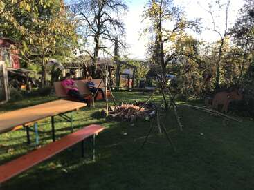 Une scène de jardin paisible avec deux bancs, un foyer, des structures en bois, des arbres et deux personnes se relaxant sur un banc sous le soleil de l'après-midi.