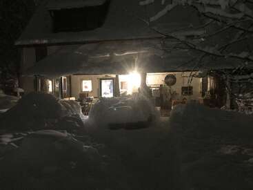 Une maison confortable brille chaleureusement dans la nuit, entourée de profondes congères et d'arbres couverts de neige, évoquant une scène hivernale paisible remplie de calme et de tranquillité.