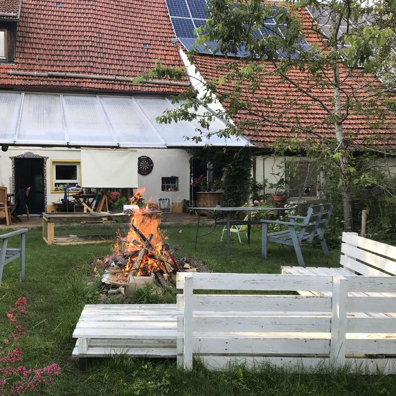 Une arrière-cour accueillante présente un feu de joie entouré de bancs en bois blanc, de sièges d'extérieur, de verdure et d'une maison avec des panneaux solaires et un toit en tuiles rouges en vue.