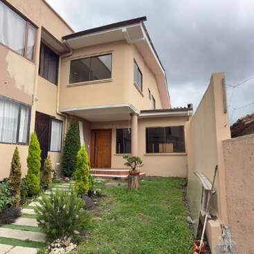 Una casa de dos plantas de color beige con grandes ventanas, una puerta marrón y un pequeño jardín delantero con arbustos y césped, bajo un cielo nublado. Cerca hay una escalera apoyada contra la pared.