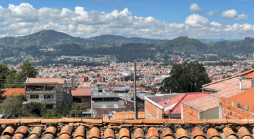 Una vista panorámica de la ciudad muestra tejados de tejas rojas en primer plano, una extensa localidad en el valle, rodeada de exuberantes montañas verdes y un cielo mayoritariamente nublado.