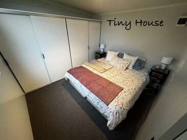 Um aconchegante quarto de uma casinha compacta tem uma cama grande com roupa de cama floral, mesinhas de cabeceira com abajures, um armário embutido com porta de correr, piso acarpetado e uma decoração de parede com o tema “Tiny House”.
