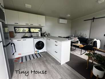 Esta imagem mostra o interior de uma pequena casa moderna com uma cozinha compacta, máquina de lavar, uma aconchegante área de estar, decoração minimalista, piso cinza e plantas de interior, destacando o uso eficiente do espaço.