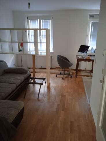 A small, tidy room with wooden flooring, a gray sofa, modern office chair, desk with computer, white shelf divider, two windows, and minimalist decor.