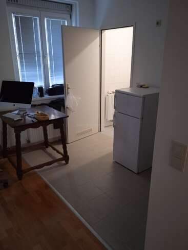 A small room with a wooden table, computer, fridge, and open door leading to a bright-tiled area. Window blinds are closed, lighting is dim.