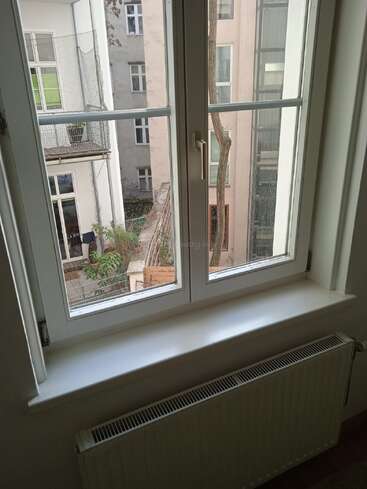 A view through a white-framed window shows an urban courtyard with apartment buildings, balconies, potted plants, a tree, and a radiator below the window.