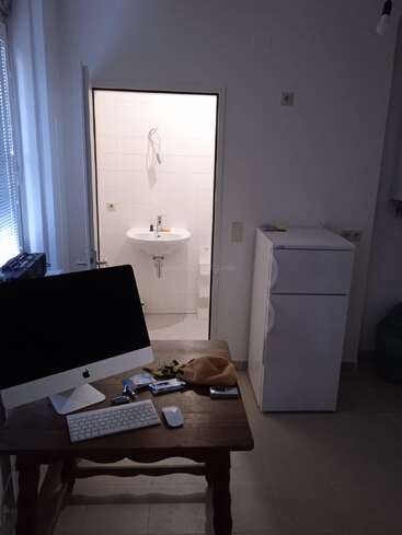 A small room with a computer on a wooden desk, various items scattered, a fridge nearby, and a view into a tiled bathroom with a sink.