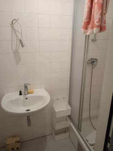 This is a small bathroom with white tiles, a wall-mounted sink, a bar of soap, a shower with a curtain, and a three-tiered storage basket.