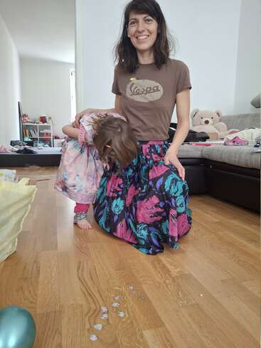 A smiling woman kneels beside a little girl in a colorful dress. The child bends forward. Toys, confetti, and a teddy bear are visible behind them.