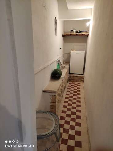 Ein schmaler Raum mit rot-weiß kariertem Boden, einer Wassermelone auf einem Steinregal, einem kleinen Kühlschrank, Küchenutensilien auf einem Holzregal und weißen Wänden.