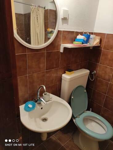 Das ist ein kleines Badezimmer mit braunen Wandfliesen, einem runden Spiegel, einem weißen Waschbecken, einem hellblauen Toilettensitz, Pflegeprodukten auf einem Regal und einem Duschvorhang.