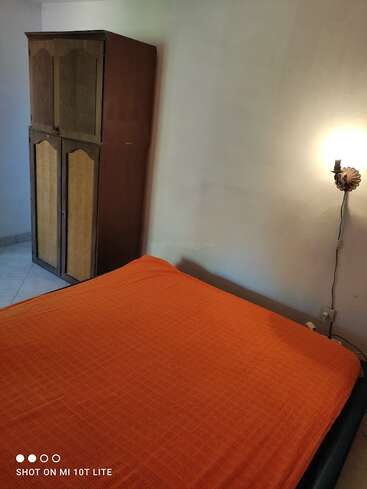 Das Bild zeigt ein schlichtes Schlafzimmer mit einer orangefarbenen Tagesdecke, einem Kleiderschrank aus Holz in der Ecke und einer Wandlampe, die für sanftes Licht sorgt.