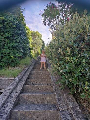 Ein kleines Kind steht auf einer Steintreppe, umgeben von üppig grünen Büschen und Bäumen, unter strahlend blauem Himmel, hält etwas in der Hand und schaut nach unten. Eine friedliche Szene.