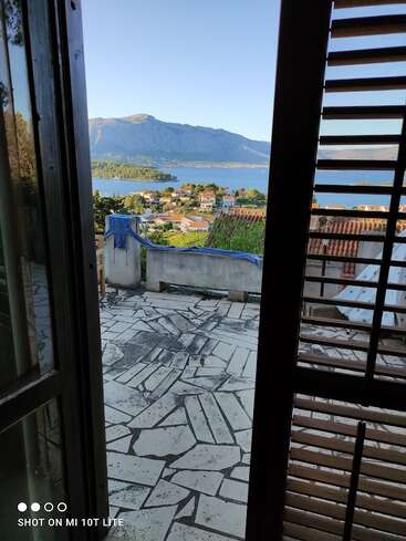 Das Foto, das durch ein offenes Fenster mit Fensterläden aufgenommen wurde, zeigt eine geflieste Terrasse, Dächer, ein malerisches Küstendorf, üppiges Grün und majestätische Berge unter blauem Himmel.
