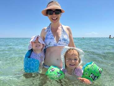 Uma mulher e duas crianças pequenas estão curtindo alegremente um dia ensolarado na praia, com os pés na água cristalina, todos sorridentes e usando chapéus e boias.