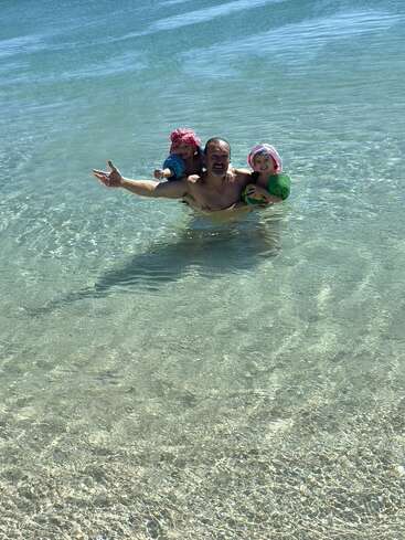 Un homme se tient joyeusement dans l'eau peu profonde et limpide de la plage avec deux jeunes enfants, tous deux coiffés d'un chapeau et équipés de brassards, profitant ensemble d'une journée ensoleillée et pleine de joie.