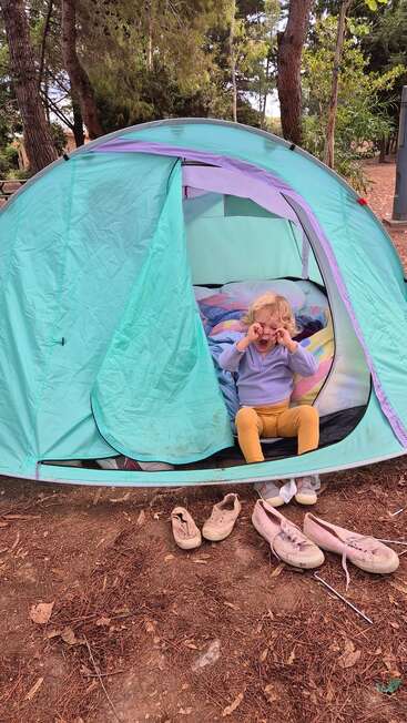 Un jeune enfant est assis à l'entrée d'une tente turquoise et se frotte les yeux. Plusieurs paires de chaussures sont posées à l'extérieur. La scène se déroule dans une forêt.