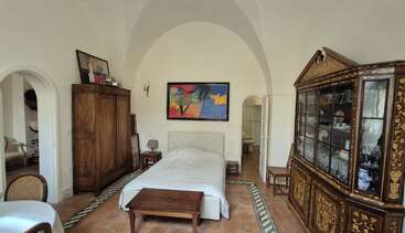 Cette élégante chambre à coucher comprend un lit double, une armoire en bois, une vitrine décorée, des œuvres d'art abstrait colorées, un sol carrelé, des plafonds voûtés, un éclairage chaleureux et une salle de bains partiellement visible.