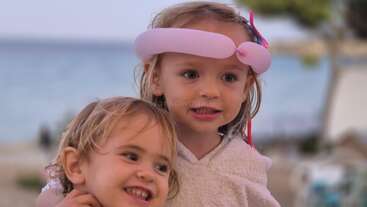 Deux jeunes enfants sourient et s'étreignent au bord de la plage. L'un d'eux porte un chapeau en forme de ballon rose. L'arrière-plan est flou, la mer et les arbres apparaissant légèrement estompés.