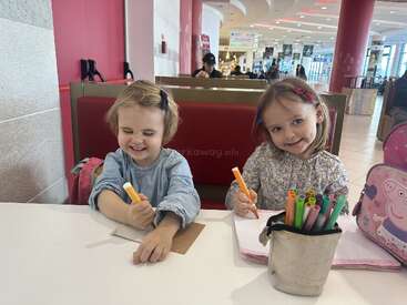 Deux petites filles sont assises à une table dans un café lumineux ; elles s'amusent à dessiner ensemble avec des feutres de couleur. Elles sourient toutes les deux, baignées dans une atmosphère joyeuse et créative.
