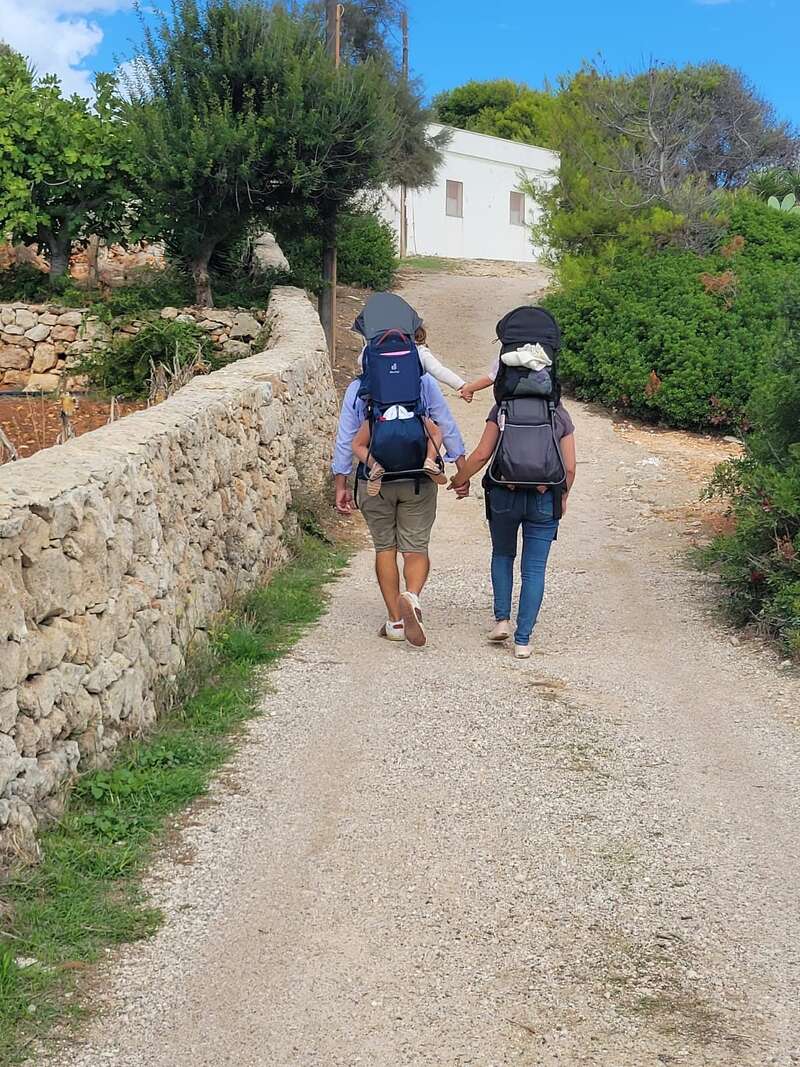 Un couple marche main dans la main sur un sentier champêtre, chacun portant un enfant dans un porte-bébé dorsal, au milieu de la verdure et des murs de pierre.