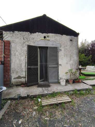 A small, weathered building with peeling paint and open metal shutters. Several potted plants sit outside, and the ground is rough with scattered debris and wood planks.