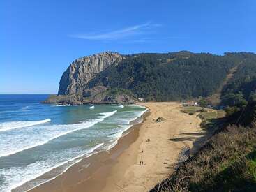A praia de areia dourada se estende ao longo da costa, com as ondas batendo suavemente. Montanhas cobertas de florestas verdes se erguem ao fundo, sob um céu azul vibrante. Poucas pessoas apreciam a paisagem serena.