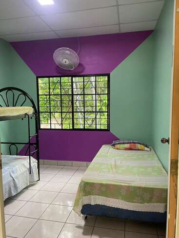 Dieses kleine, gepflegte Zimmer verfügt über ein Einzelbett, ein Etagenbett, pastellgrüne Wände mit violetten Akzenten, einen Deckenventilator, einen Fliesenboden und ein Fenster.