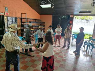 Ocho adultos están de pie formando un círculo en una habitación con baldosas a cuadros; algunos se dan la mano. Parece que están participando en una actividad en grupo o en un taller.