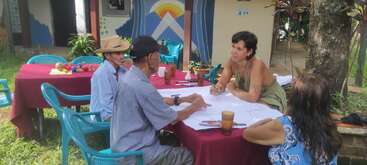Cuatro personas están sentadas alrededor de una mesa al aire libre, conversando y tomando notas en grandes hojas de papel. El ambiente es distendido, con árboles y sillas de colores.