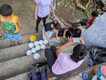 Un grupo de niños y adultos se reúne en unas escaleras al aire libre, preparando pintura en cuencos. Una persona hace una foto mientras los demás mezclan colores, rodeados de materiales.