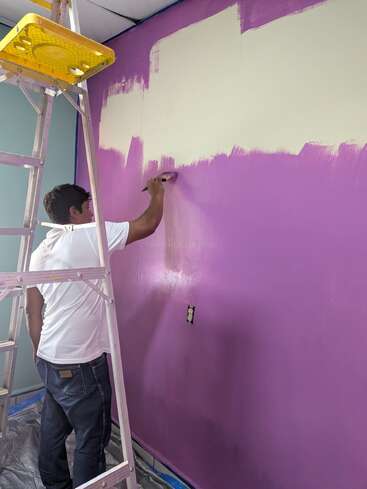 Una persona con una camisa blanca está pintando una pared morada de color beige con un pincel. Hay una escalera cerca y el suelo está cubierto con plástico protector.