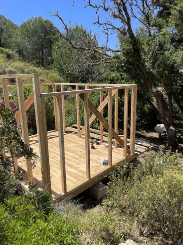 A small wooden structure is being framed in a forested, sunlit area. Only the floor and wall frames are built; tools and supplies are on the floor.
