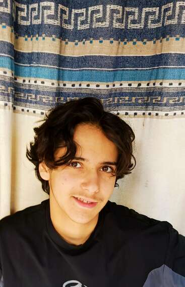Un jeune homme aux cheveux bruns ondulés, vêtu d'un t-shirt noir, sourit doucement à la caméra. Derrière lui se trouve un rideau à motifs bleus et beiges.