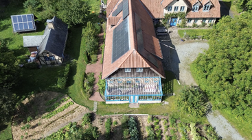 This aerial image shows a rural property with solar panels on roofs, a large main house, garden beds, trees, greenhouses, and additional small structures surrounded by greenery.