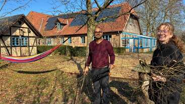 A man and woman happily gather sticks in a sunny yard. Behind them is a charming house with solar panels, blue windows, and a colorful hammock.