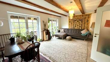 This cozy living room features wooden beams, large windows, a gray sectional sofa, a decorative rug, plants, a chandelier, and a tapestry accent on the wall.