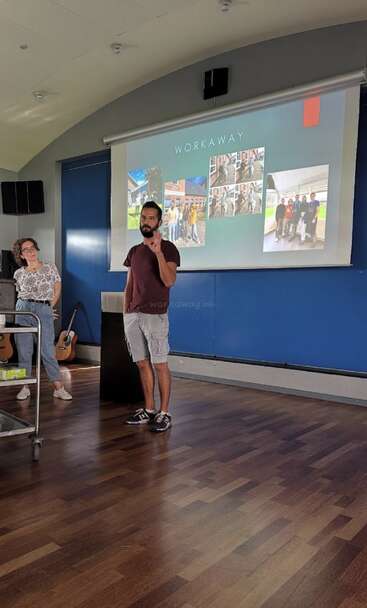 Un hombre presenta delante de una pantalla proyectada "WORKAWAY" con fotos. Una mujer está cerca, observando. Se encuentran en una moderna sala de paredes azules.