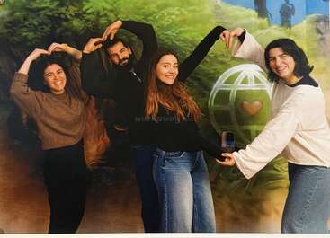 Four smiling people stand in front of a colorful mural, posing together to form the word "LOVE" with their arms, expressing friendship, happiness, and creativity.