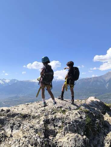 Zwei junge Wanderer, mit Helmen und Rucksäcken, stehen auf einem felsigen Berggipfel und blicken unter strahlend blauem Himmel auf ein atemberaubendes Tal und schneebedeckte Berge.